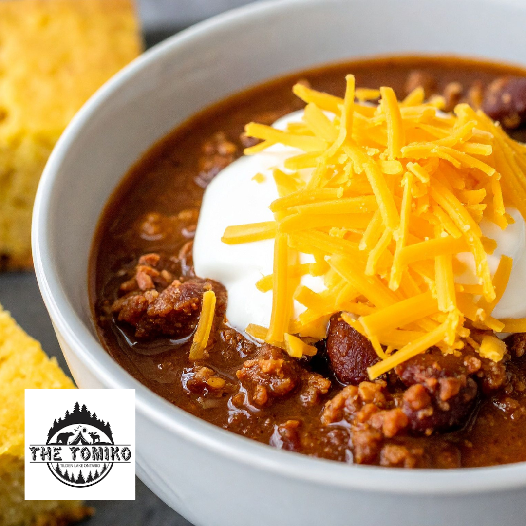 Bowl of chili with sour cream and shredded cheese, featuring The Tomiko logo.