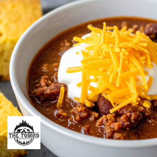 Bowl of chili with sour cream and shredded cheese, featuring The Tomiko logo.