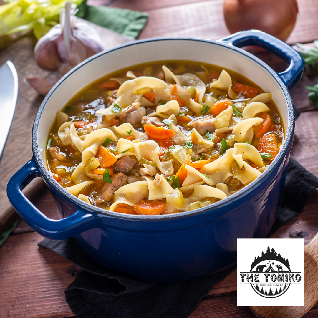 Blue pot of noodle soup with vegetables on a wooden table, featuring The Tomiko logo.