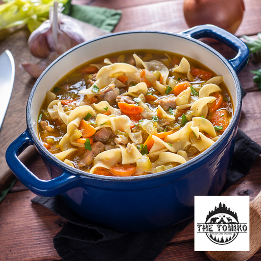 Blue pot of noodle soup with vegetables on a wooden table, featuring The Tomiko logo.