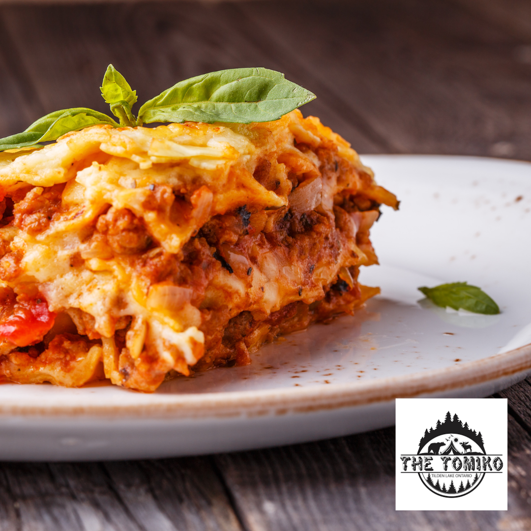 Slice of lasagna on a plate with basil leaves, featuring 'The Tomoko' logo.